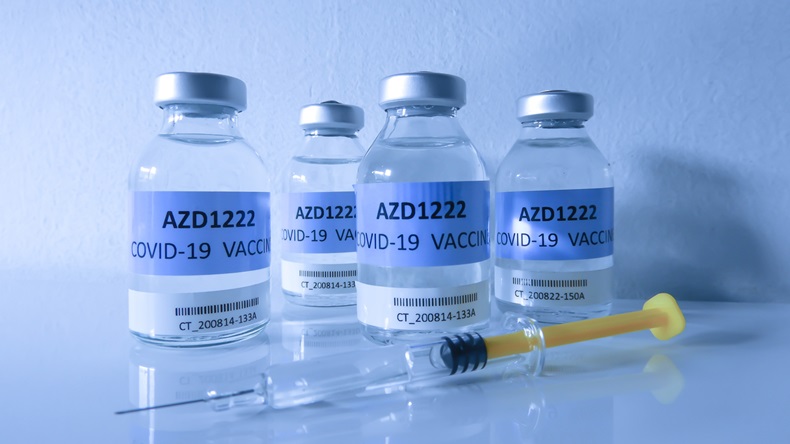 Four transparent glass vials with a test COVID-19 vaccine, named AZD1222, and a glass syringe with yellow piston on a blue surface and with blue background