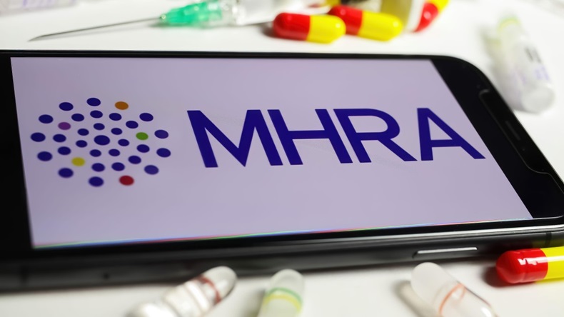 Closeup of mobile phone screen with logo lettering of UK MHRA agency, serum vials and syringe background - Image ID: 2H94BWR