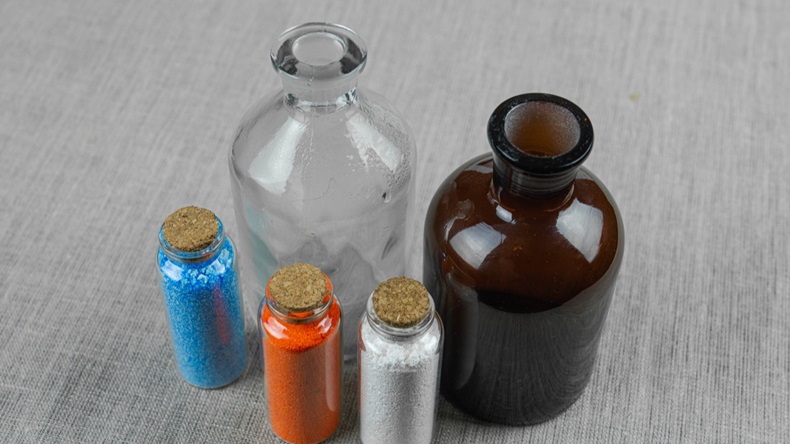 Red, white and blue powder and crystals in small bottle on gray linen background near two big transparent and dark bottles