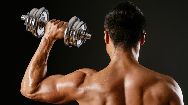 Photo of an Asian male exercising with dumbbells and doing a single shoulder press over dark background.